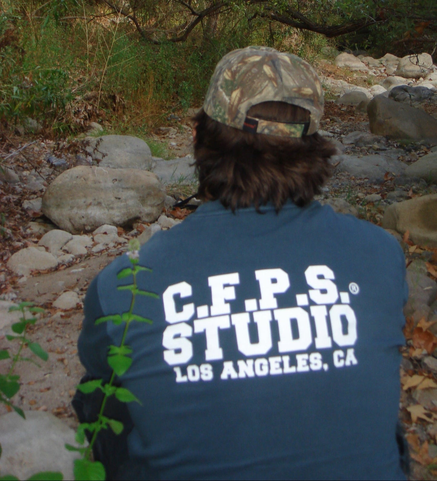 Person wearing a blue jacket with 'C.F.P.S. Studio Los Angeles, CA' on the back, standing in a forested area.