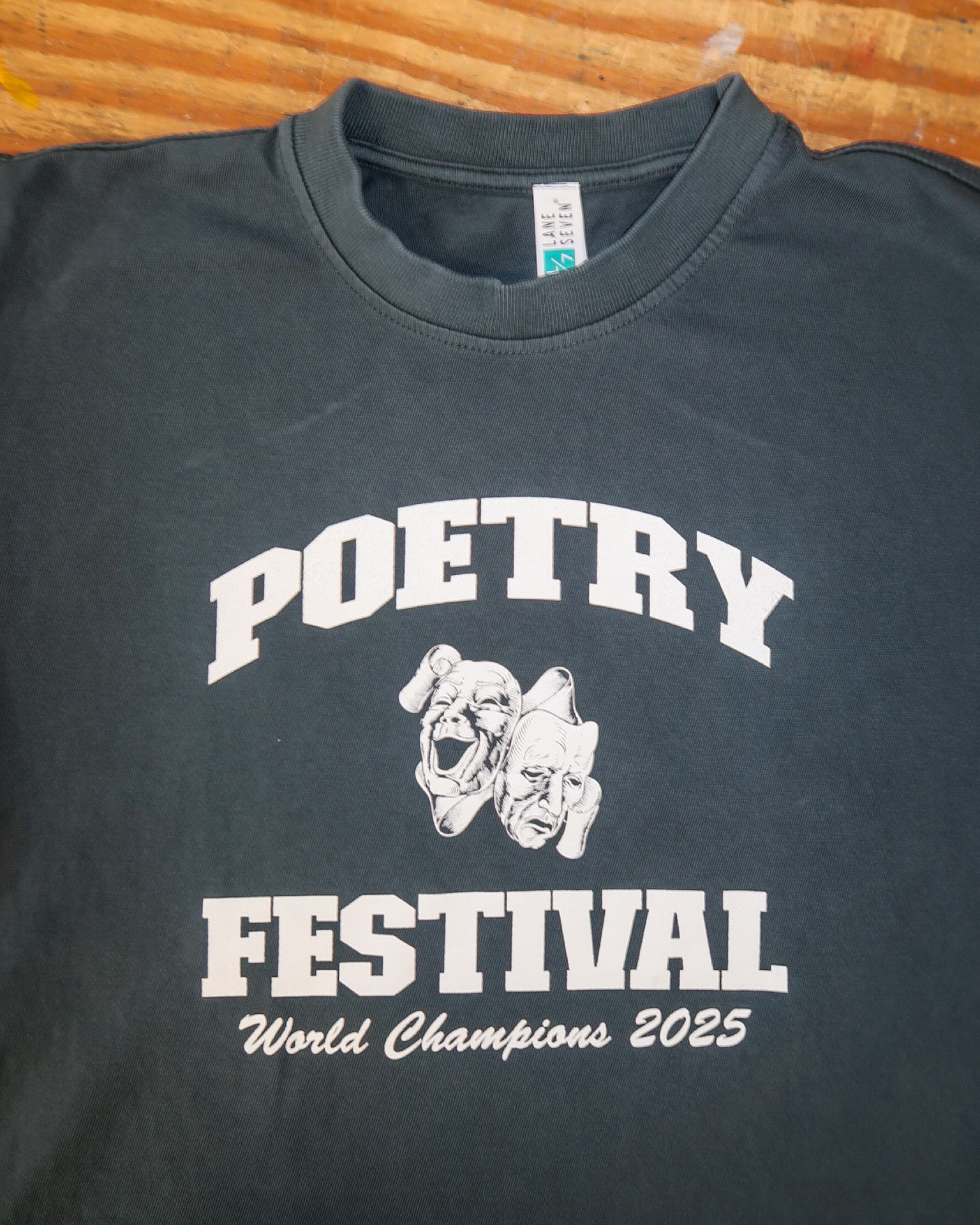 Navy t-shirt with 'POETRY FESTIVAL World Champions 2025' text and graphic on a wooden surface