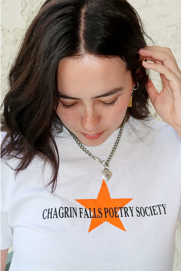 A woman with dark hair looking down, wearing a white t-shirt with a black text print 'CHAGRIN FALLS POETRY SOCIETY' and an orange star graphic, along with a silver chain necklace.